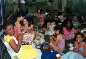 Large group of elementary students knitting together. They are smiling, sitting on the floor of a school hallway.