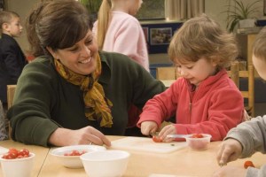 Very small child cutting vegetables with a smiling adult beside her supervising.